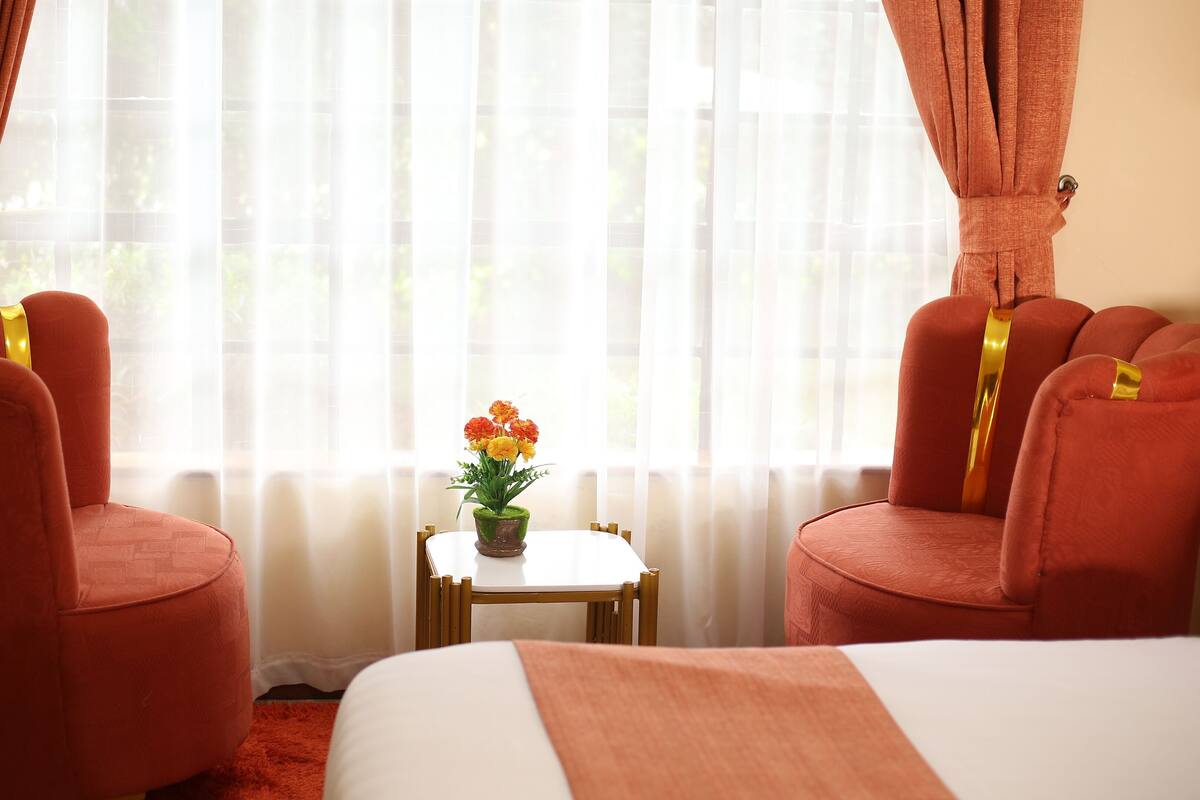 Ein Hotelzimmer mit zwei roten Polstersesseln, einem kleinen Tisch mit Topfpflanze und einem Bett mit weiß-orangener Bettdecke.