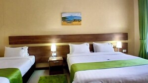 A hotel room with two beds, a headboard, bedside lamps, a green throw blanket, a painting on the wall, and a window with curtains. - LIA Hotel & Training Centre (Nairobi)