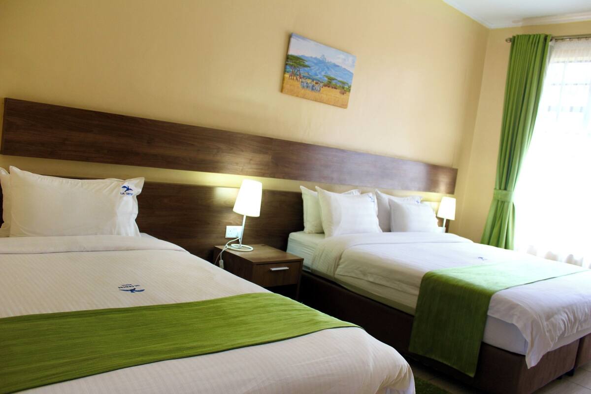 Ein Hotelzimmer mit zwei Betten, jeweils mit grüner Bettdecke, weißen Laken und einem Holz-Kopfteil. An der Wand hängt ein Gemälde und es gibt ein Fenster mit grünen Vorhängen.
