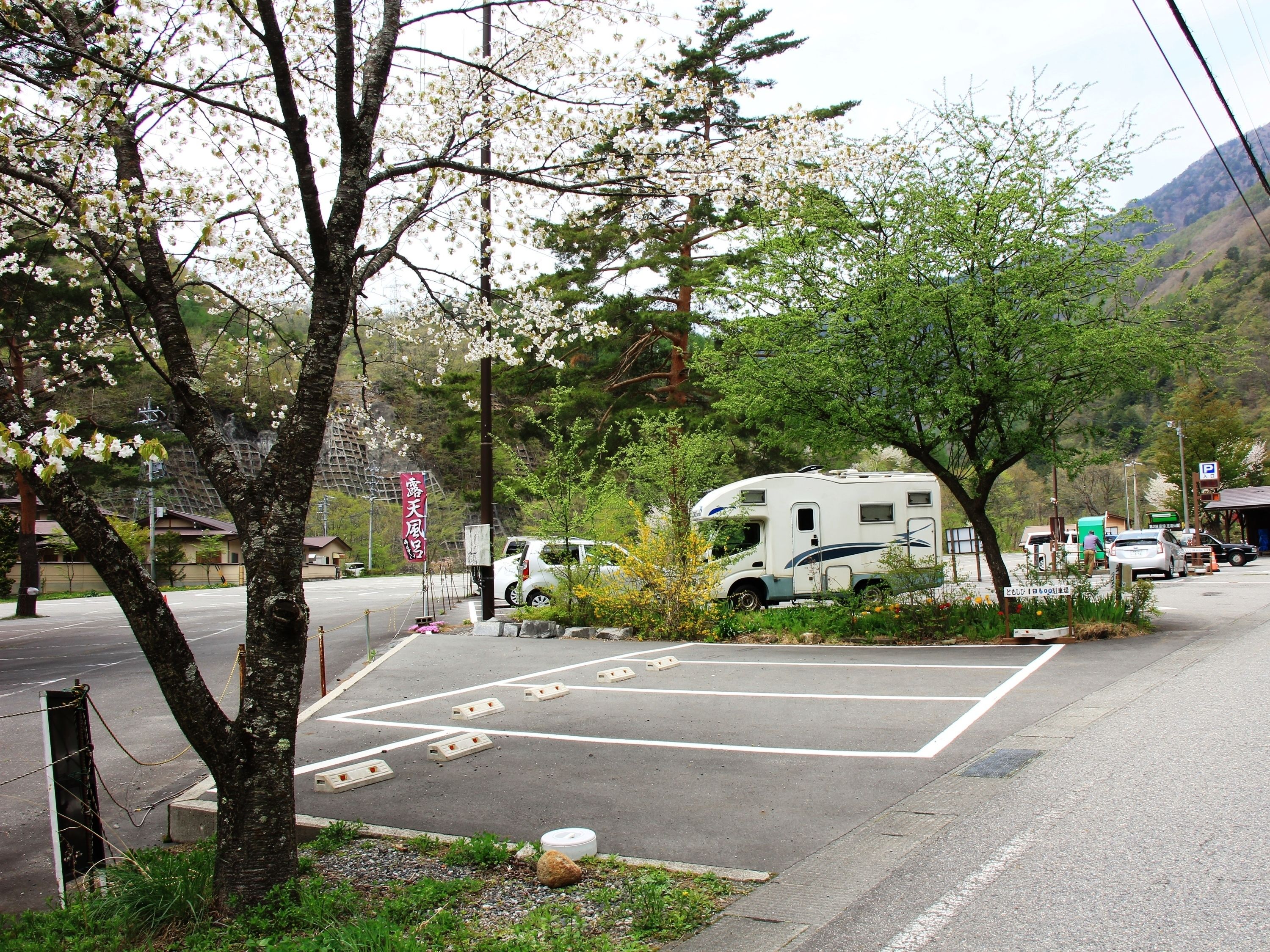 A parking area with a white RV and a car, surrounded by trees and a mountainous backdrop.