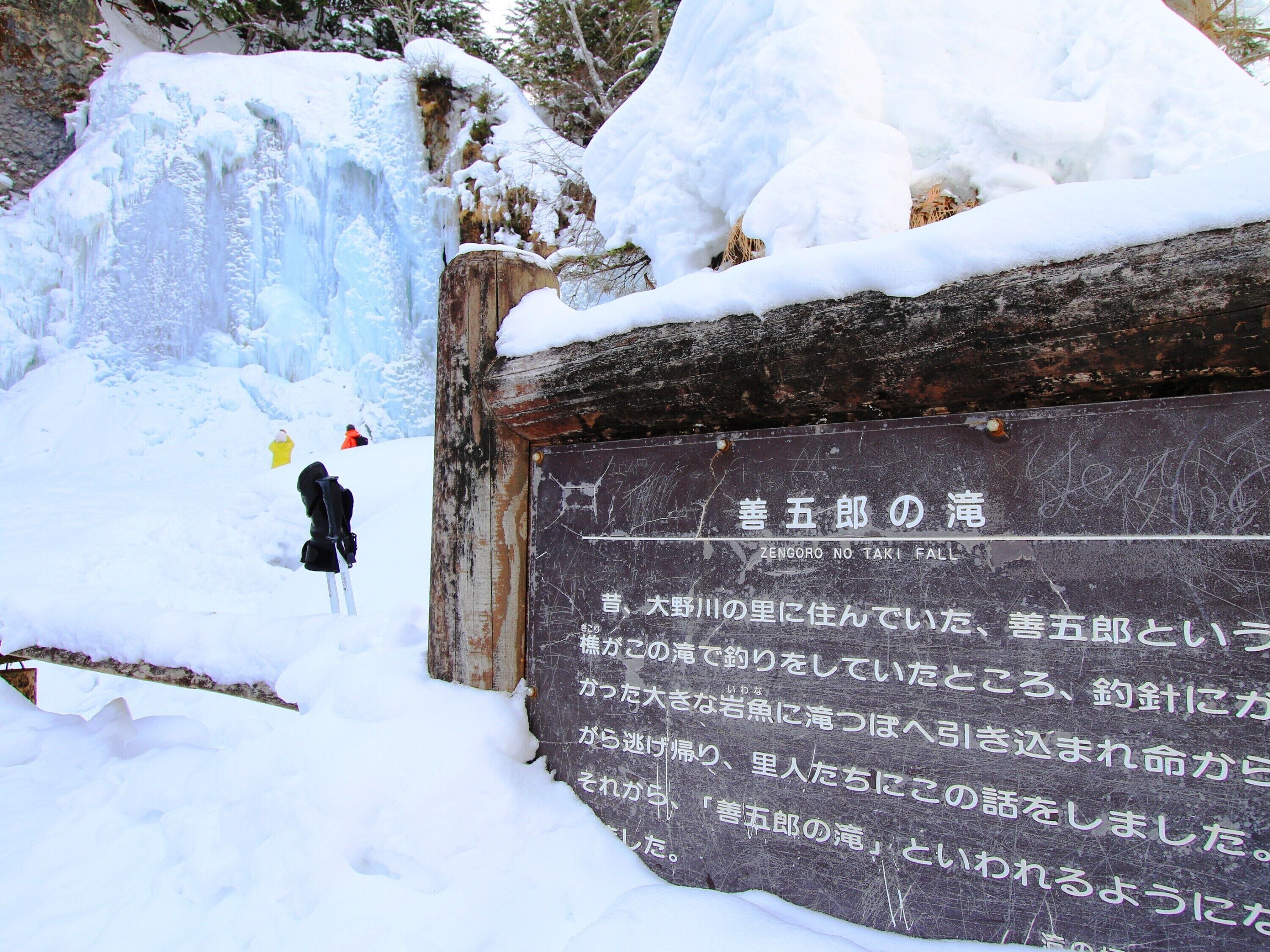 Uma placa de madeira coberta de neve com texto em japonês, indicando um local.