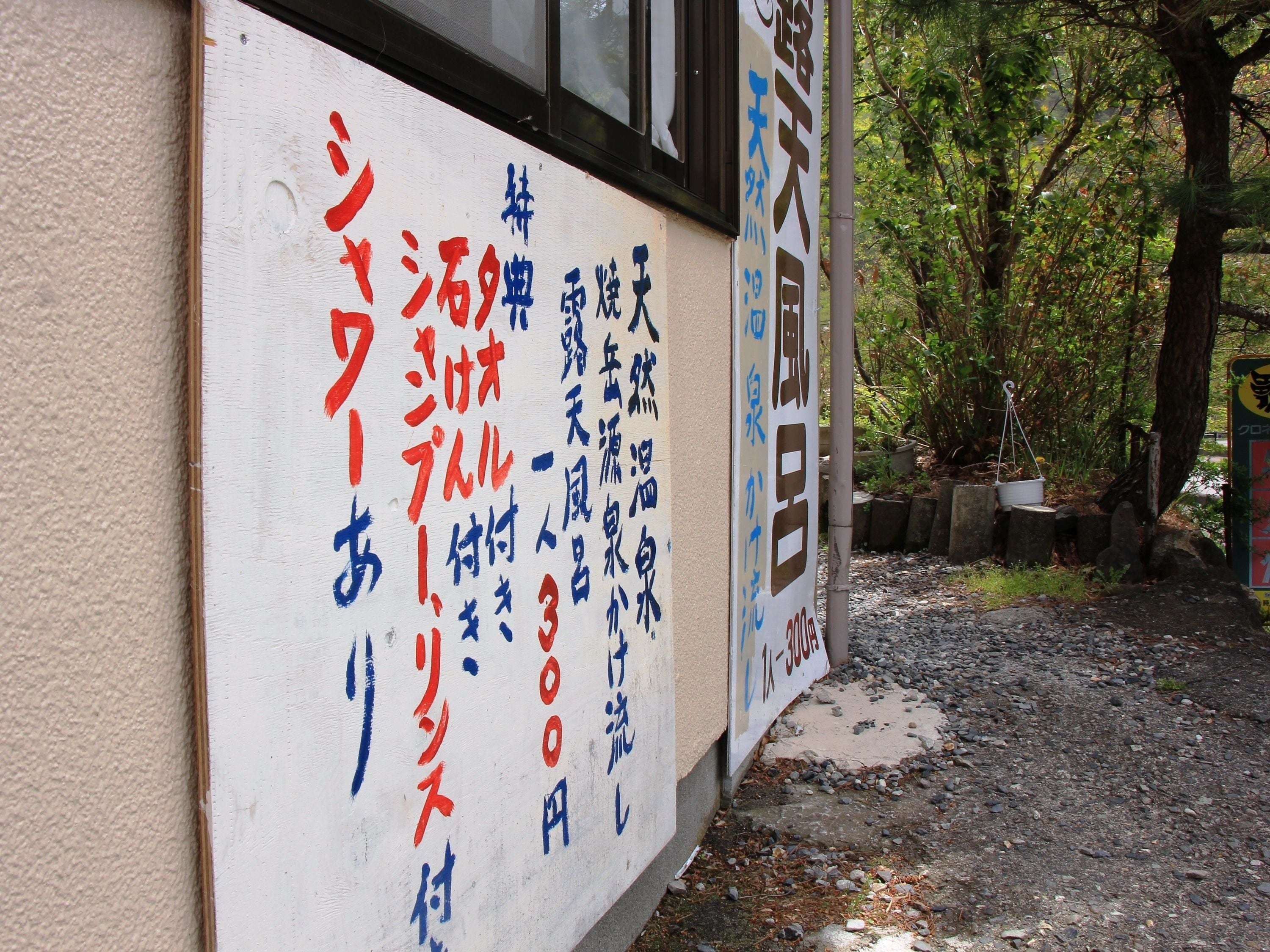 A sign with Japanese text on a building wall, surrounded by trees and gravel.