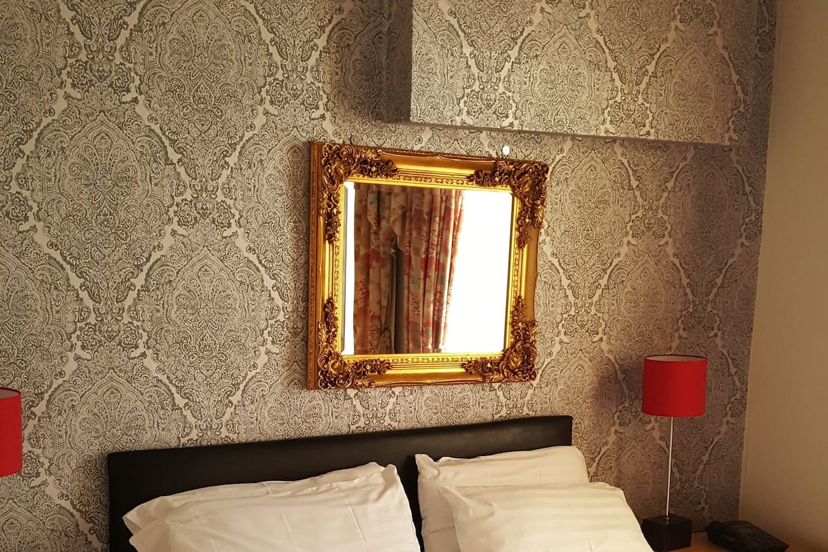 A neatly made bed with white linens, a gold-framed mirror above it, and a red lamp on a bedside table.