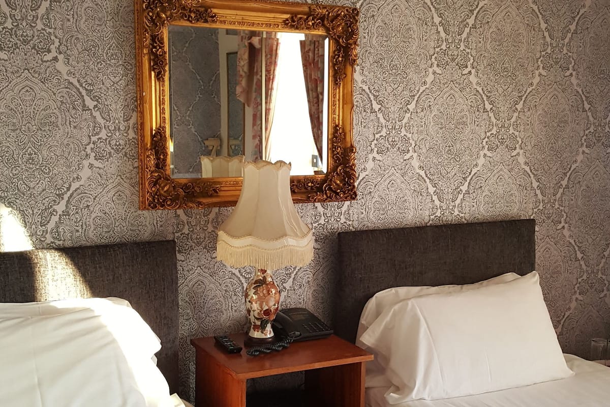 A hotel room with two beds, a nightstand with a lamp, a mirror, and patterned wallpaper.