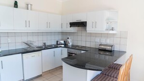 A compact kitchen with white cabinets, a black countertop, a built-in oven, and a small island with a chair. - Villa 9 (Knysna)