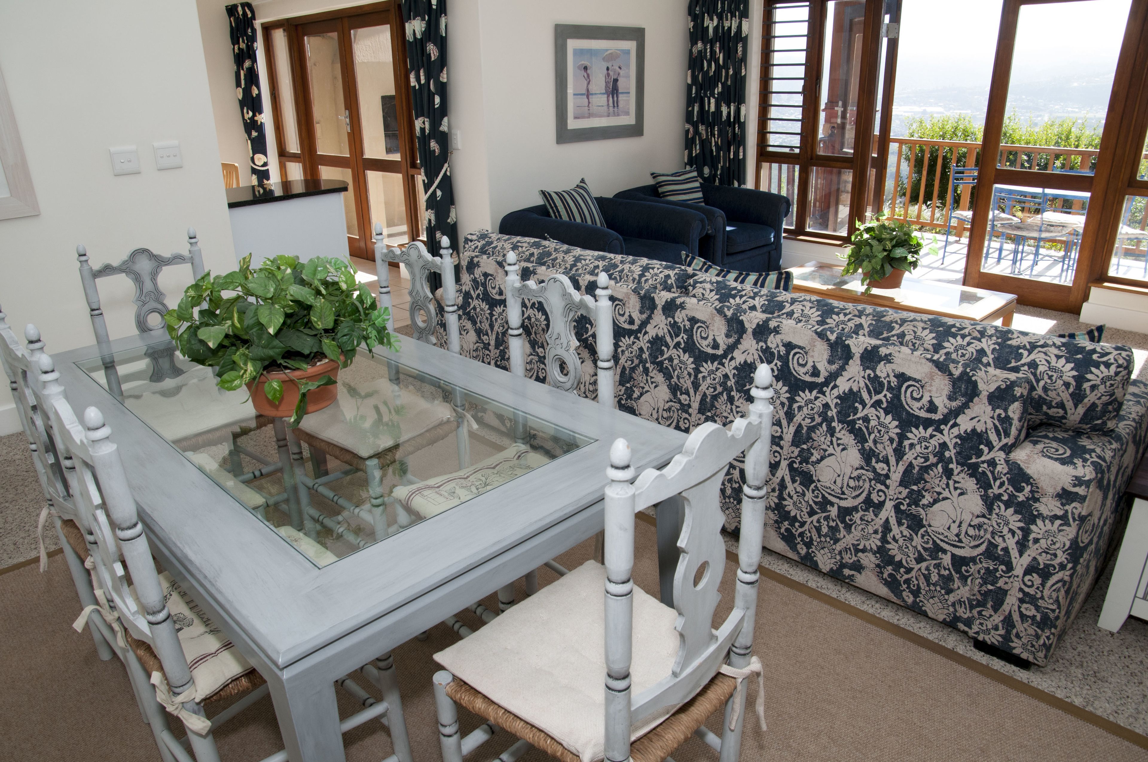 A dining area with a glass-top table, white chairs, and a blue and white patterned sofa.