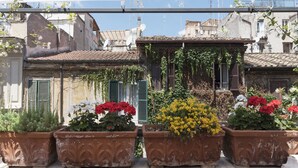 A row of terracotta planters with various flowers and plants, set against a backdrop of old buildings with tiled roofs and green shutters. - Monti Stairway to Heaven (Rome)