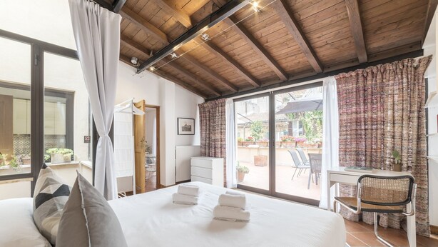 A bedroom with a large bed, a desk, and a chair. There is a window with curtains and a view of the outside. - Monti Stairway to Heaven (Rome)