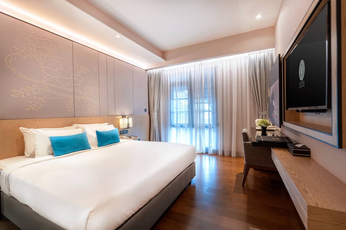 A modern hotel room with a large bed, a desk, a flat-screen TV, and a large window with curtains.