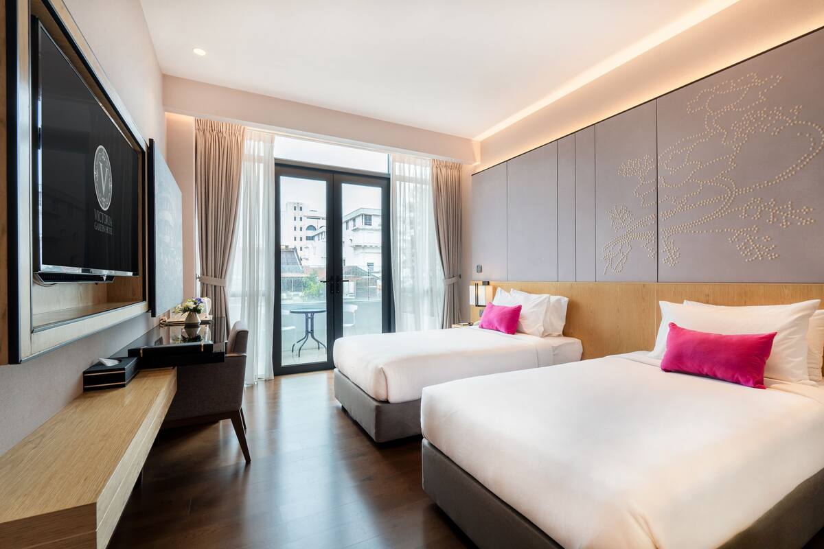 A hotel room with two beds, a TV, a desk, and a balcony with a view of buildings.