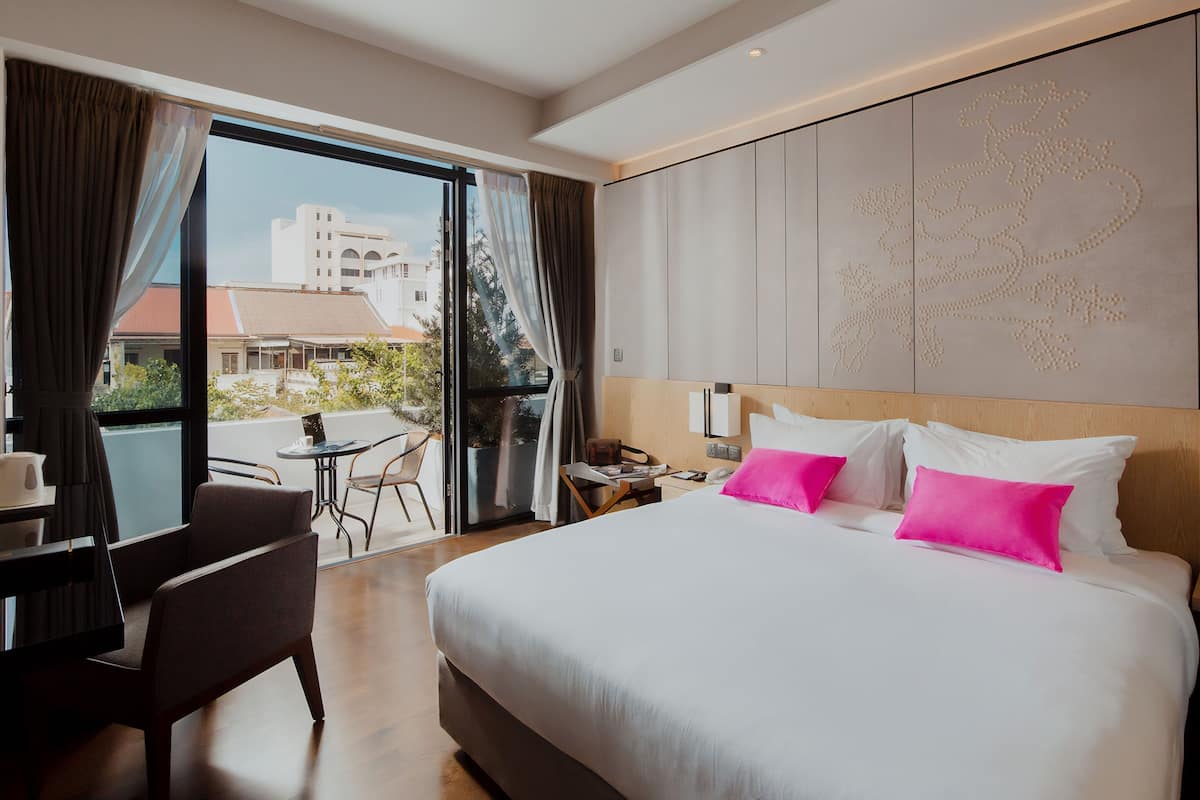 A hotel room with a large bed, a desk with a chair, a small table, and a view of buildings outside through a large window.