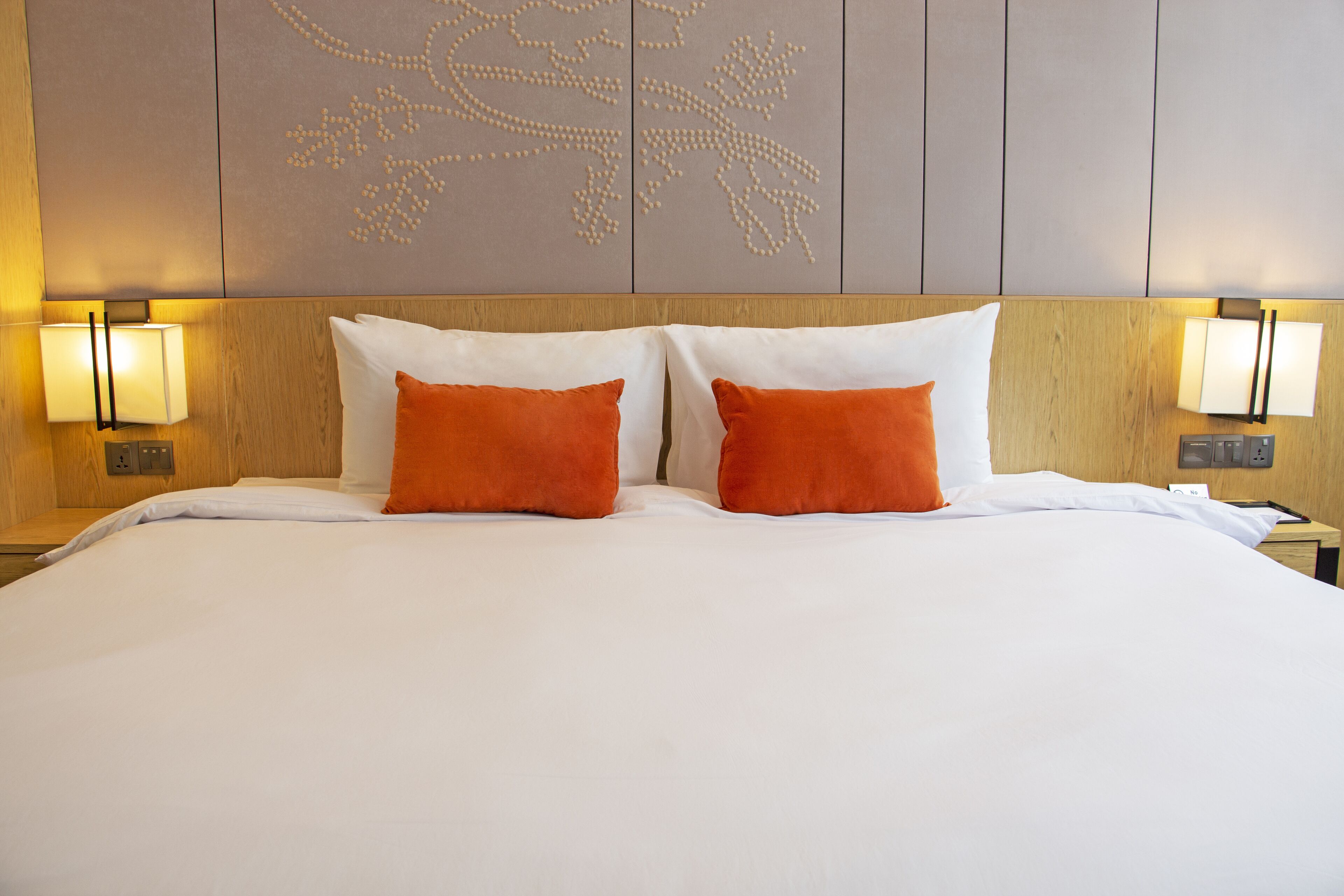 A hotel room with a large bed, two orange pillows, and a decorative wall panel.
