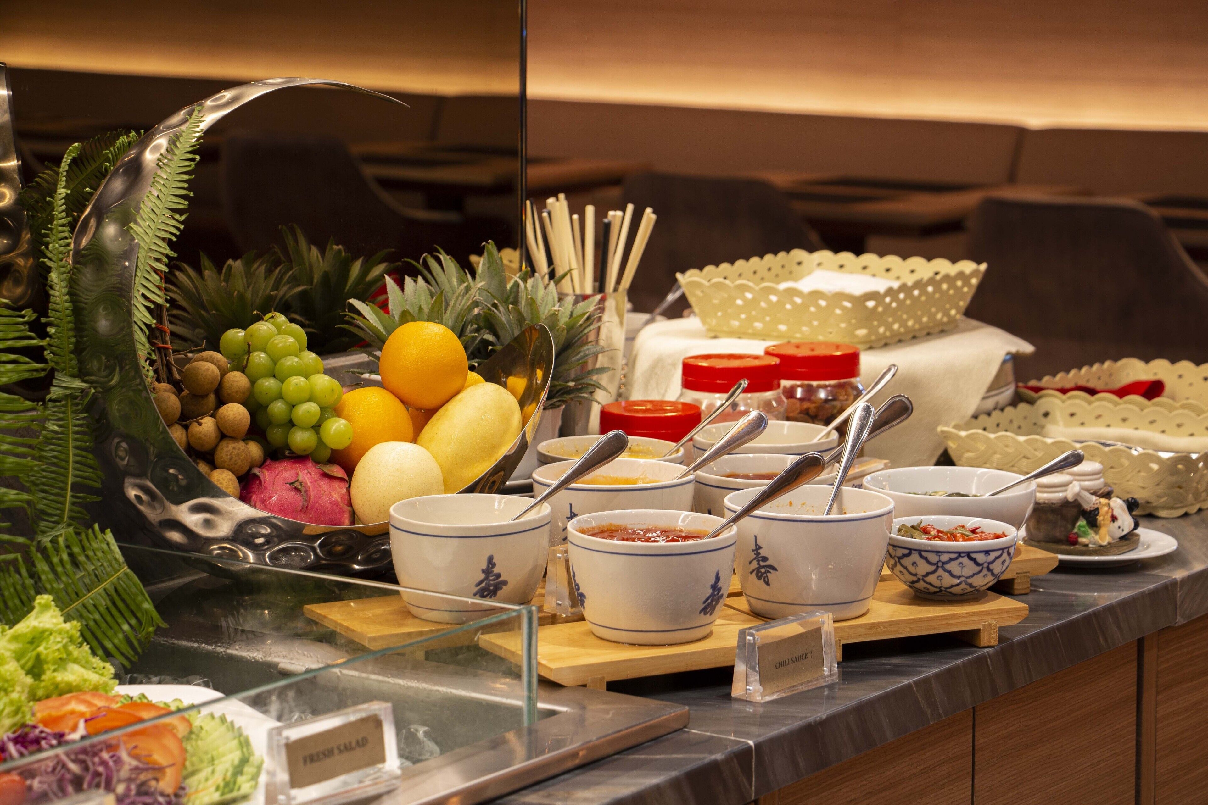 A buffet with various food items and utensils, including bowls of soup and a fruit display.