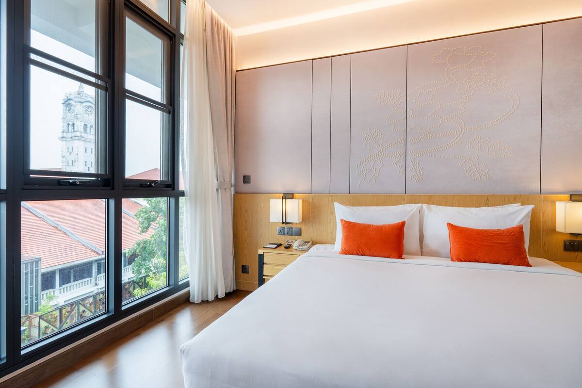 A hotel room with a large bed, two pillows, a nightstand with a lamp, and a view of a building through a window.