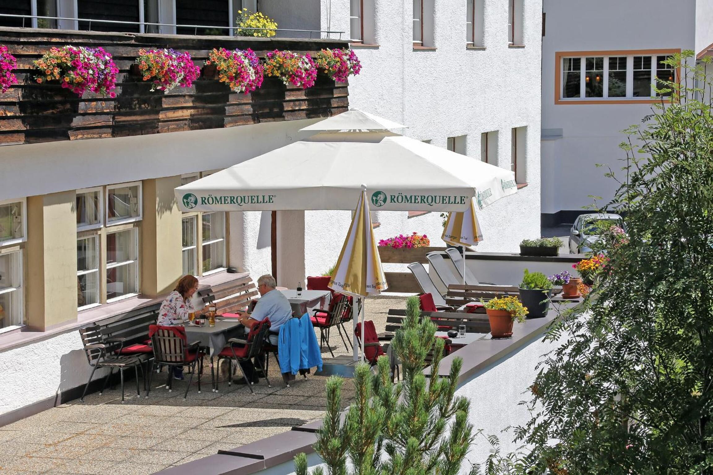 Ein Café im Freien mit Tischen, Stühlen, einem weißen Sonnenschirm und Blumenkästen.