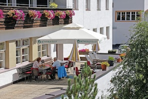 Un área de café al aire libre con mesas y sillas, una sombrilla blanca y macetas con flores.