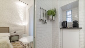 Een compacte hotelkamer met een zwart-wit geruite vloer, een bed met een zachte, witte deken, een klassieke televisie, een klein tafeltje en een kitchenette met raam.