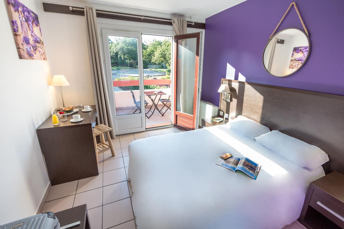 A hotel room with a bed, a desk with a lamp, a chair, a table with a glass of water, and a view of the outdoors.