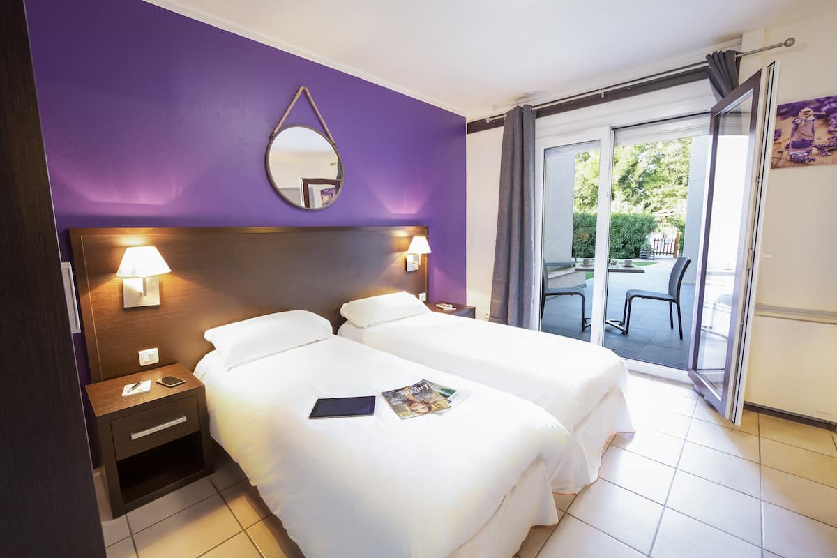 A hotel room with two beds, a nightstand with a lamp, a mirror, and a view of the outside balcony.