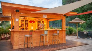 A wooden outdoor bar with a sign reading "COMMANDE AU BAR" and a "BEER" sign above the bar. There are three stools and a table with chairs.