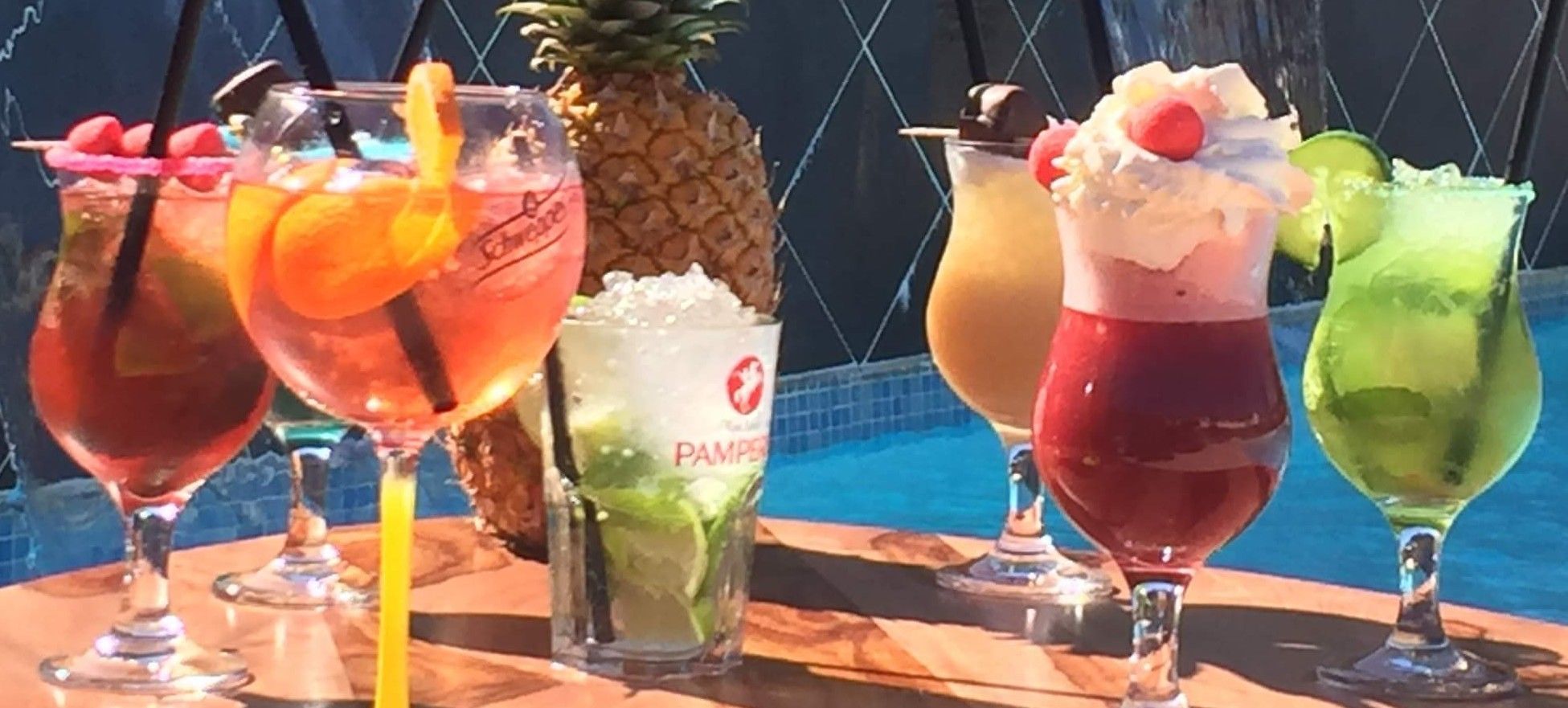 A variety of colorful cocktails on a wooden table with a pool in the background.