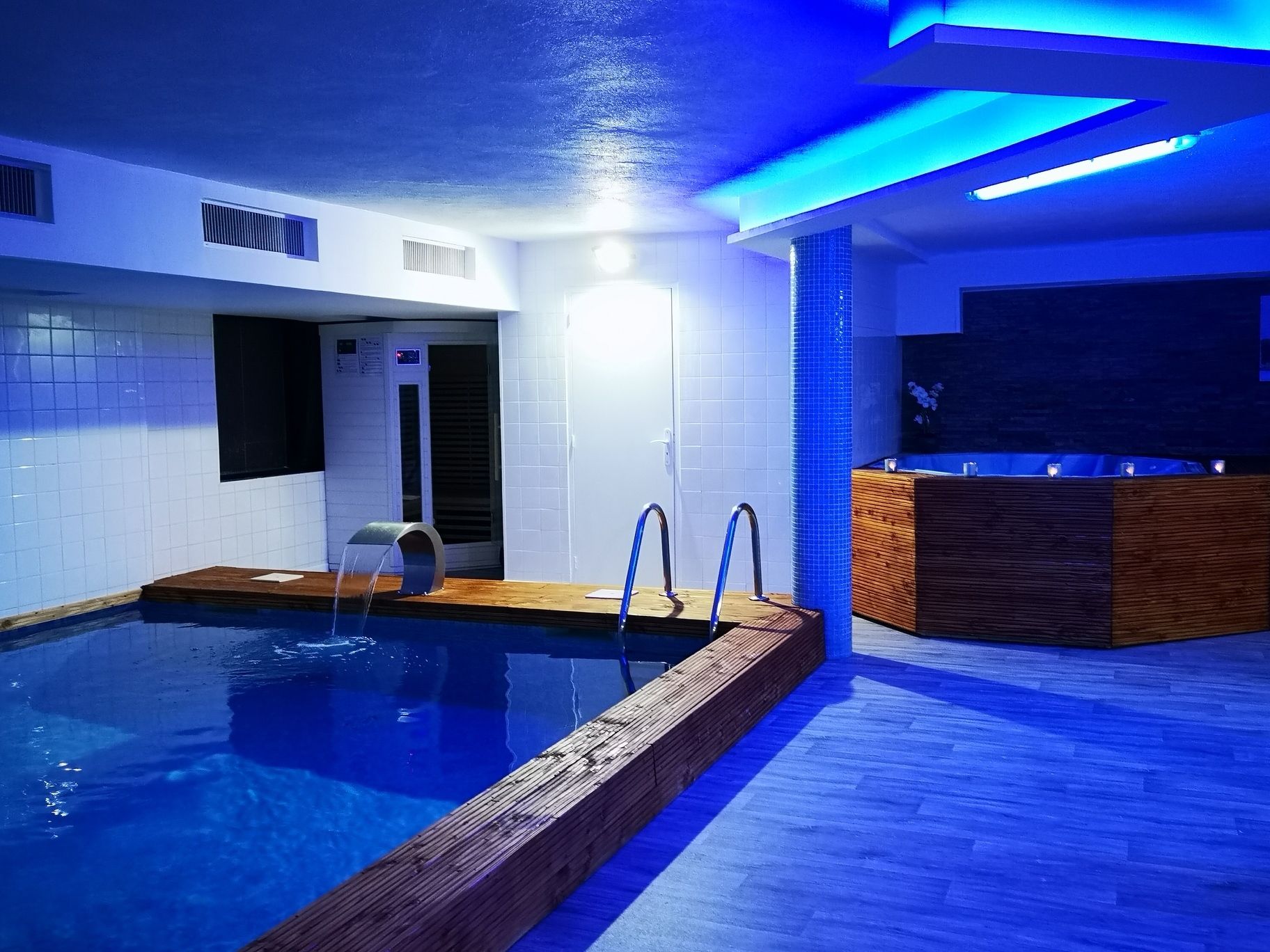 Una piscina interior con terraza de madera, zona de sauna y mostrador de recepción.
