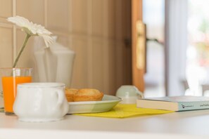 A breakfast setup with a white pitcher, a glass of orange juice, a plate with pastries, a book, and a white flower in a vase. - Casa Gaia PP (Porto Cesareo)