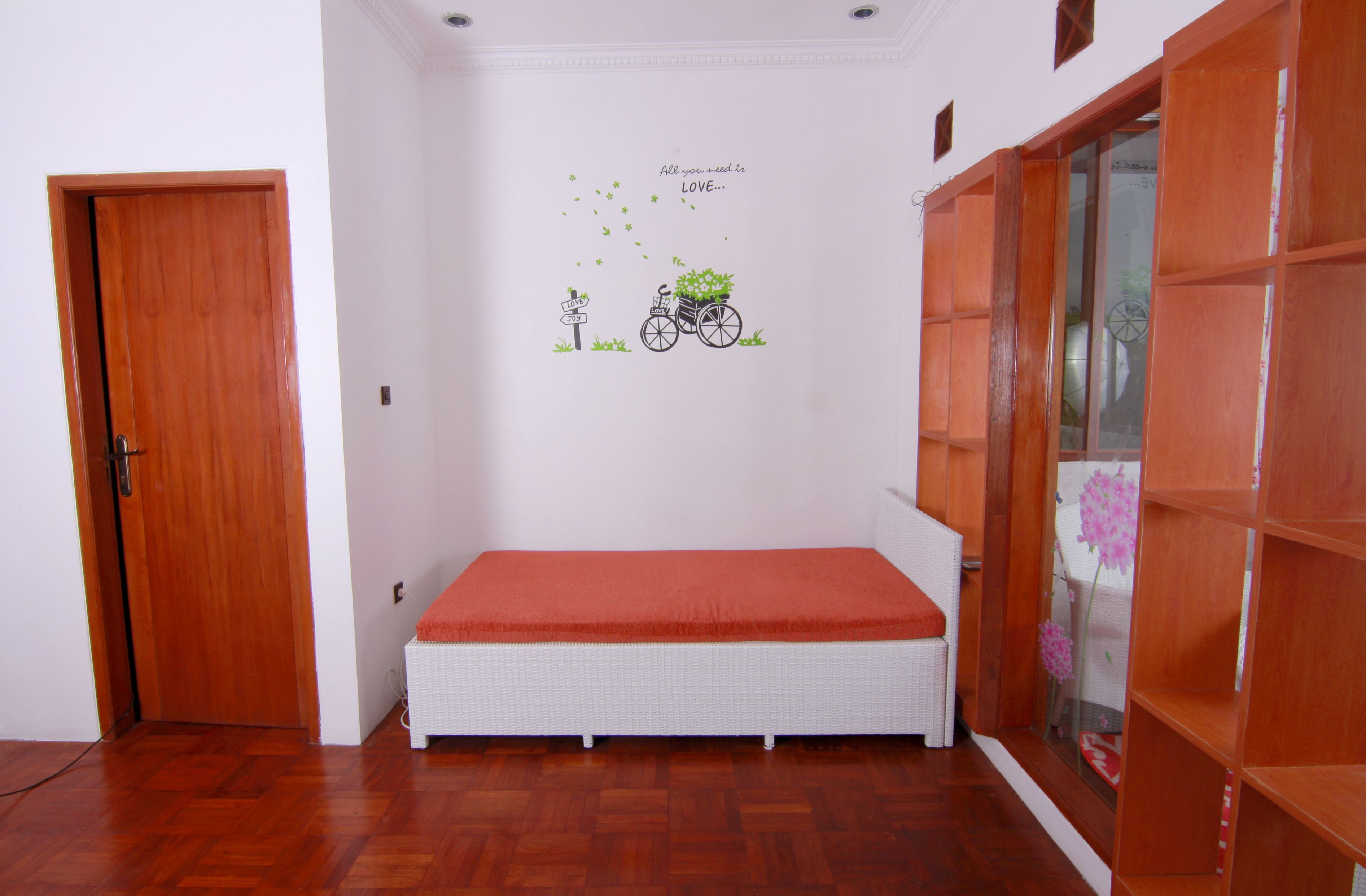 Une pièce avec une porte en bois, un lit en osier blanc recouvert d’une couverture rouge, un motif décoratif mural représentant un vélo et des fleurs, et une bibliothèque en bois.