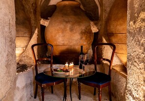 Una mesa con dos sillas, velas, una botella y copas en una habitación de paredes de piedra, con una olla grande al fondo.