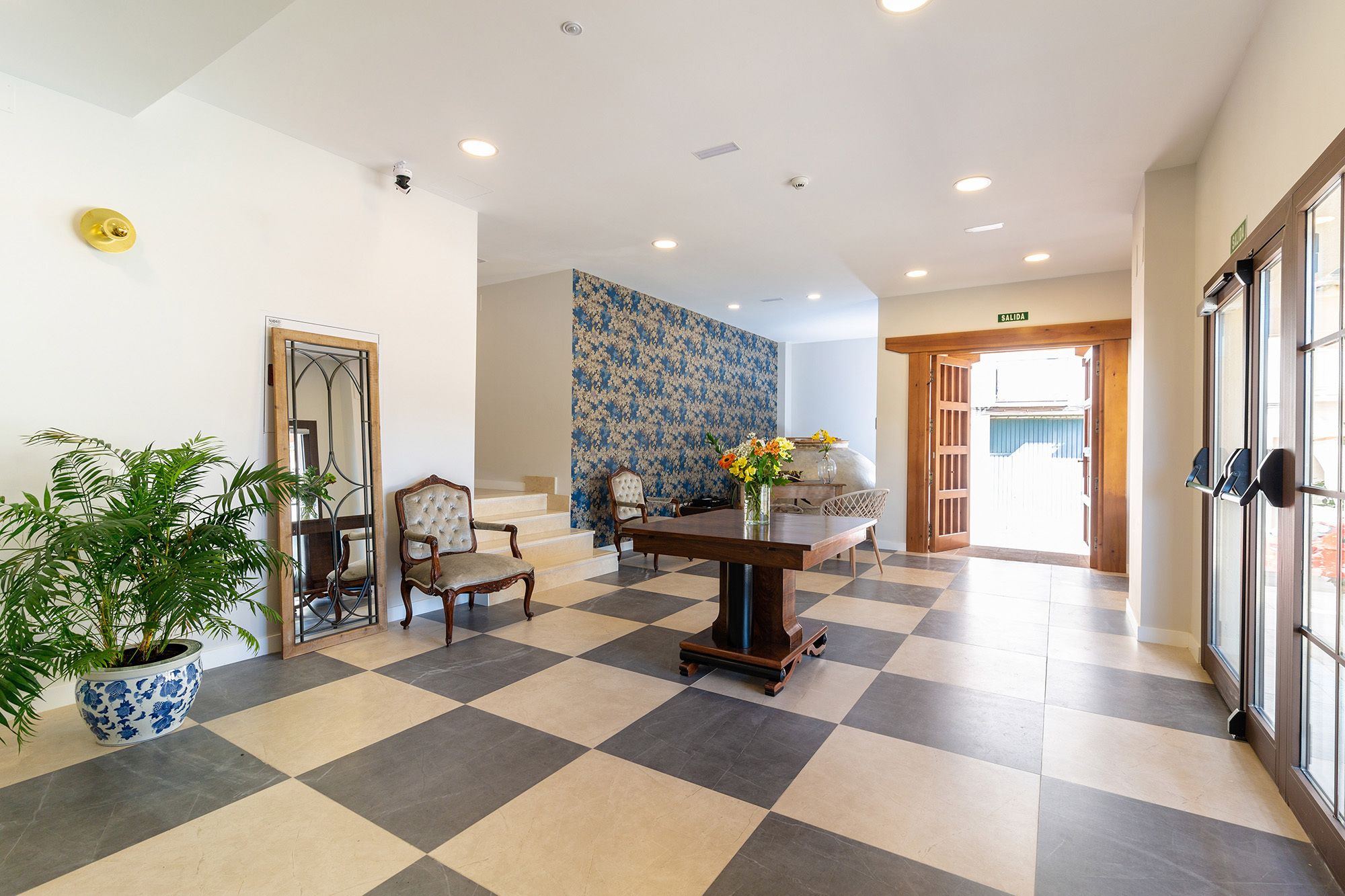 A hotel lobby with a checkered floor, a reception desk, a potted plant, and a wall with a blue floral pattern.