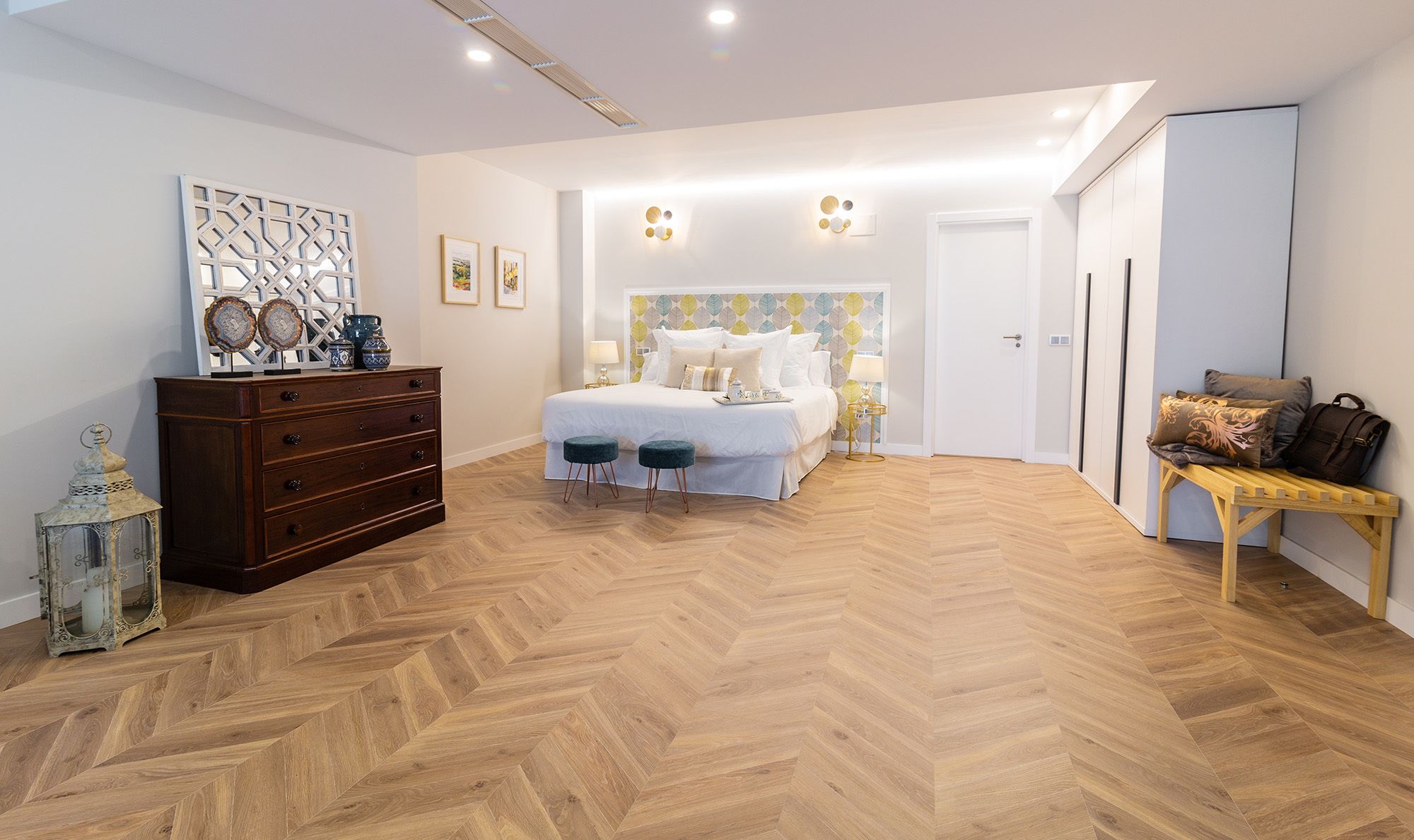 A bedroom with a herringbone wooden floor, a bed with white bedding, a dresser, and a bench with a bag.