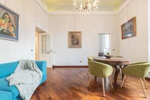 Una habitación con un sofá azul, una mesa de comedor con sillas, suelo de madera y una araña.