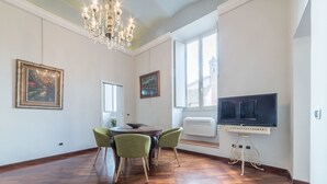 Un comedor con una mesa de madera, dos sillas verdes, una araña y un televisor sobre un mueble.