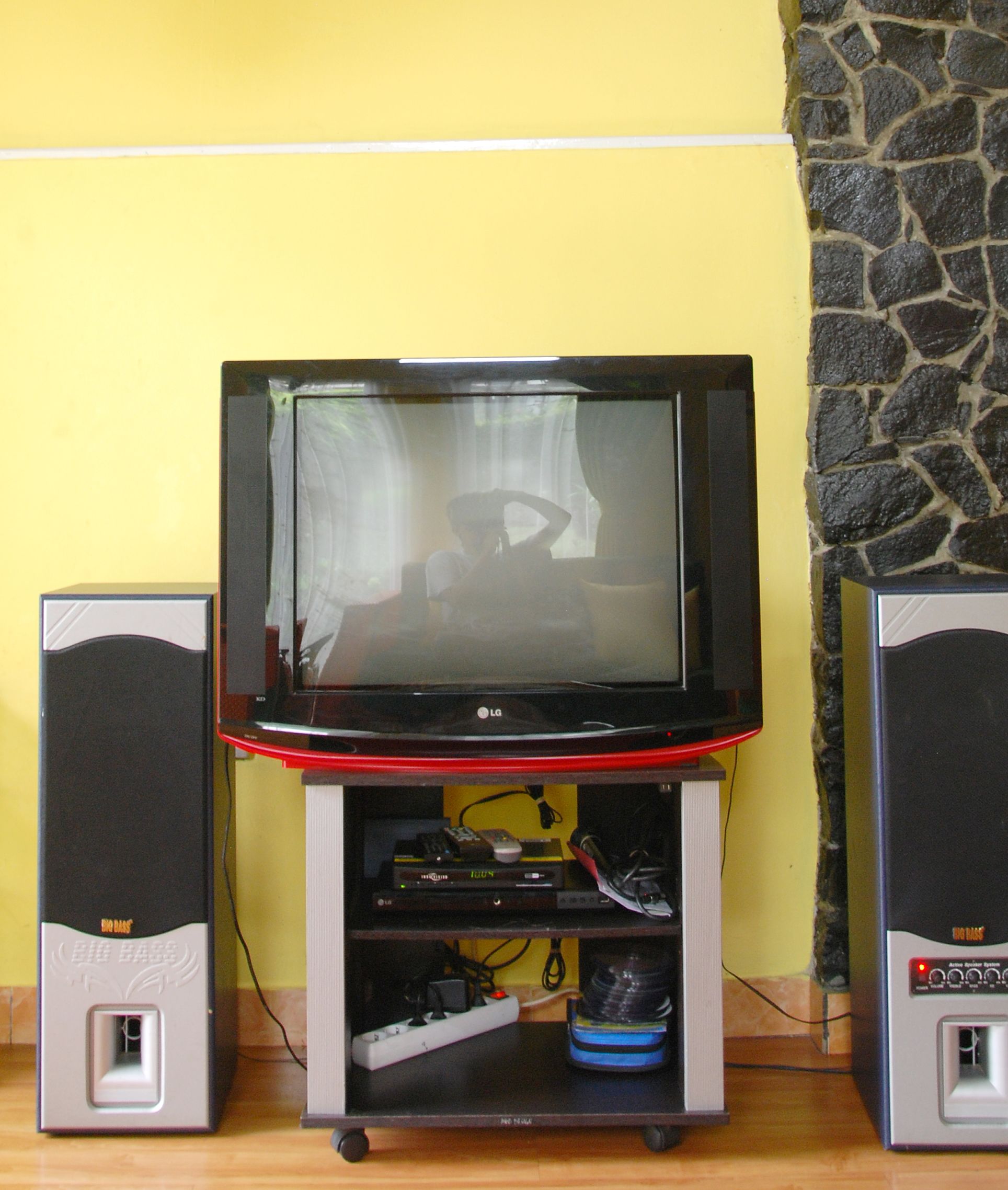A television set on a wooden stand with two speakers on either side, against a yellow wall with a stone accent.