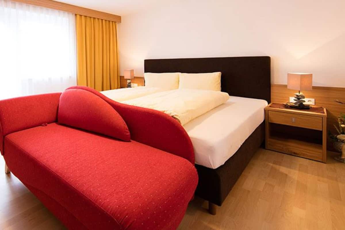 A hotel room with a bed, a red sofa, a TV, and a wooden nightstand with a lamp.