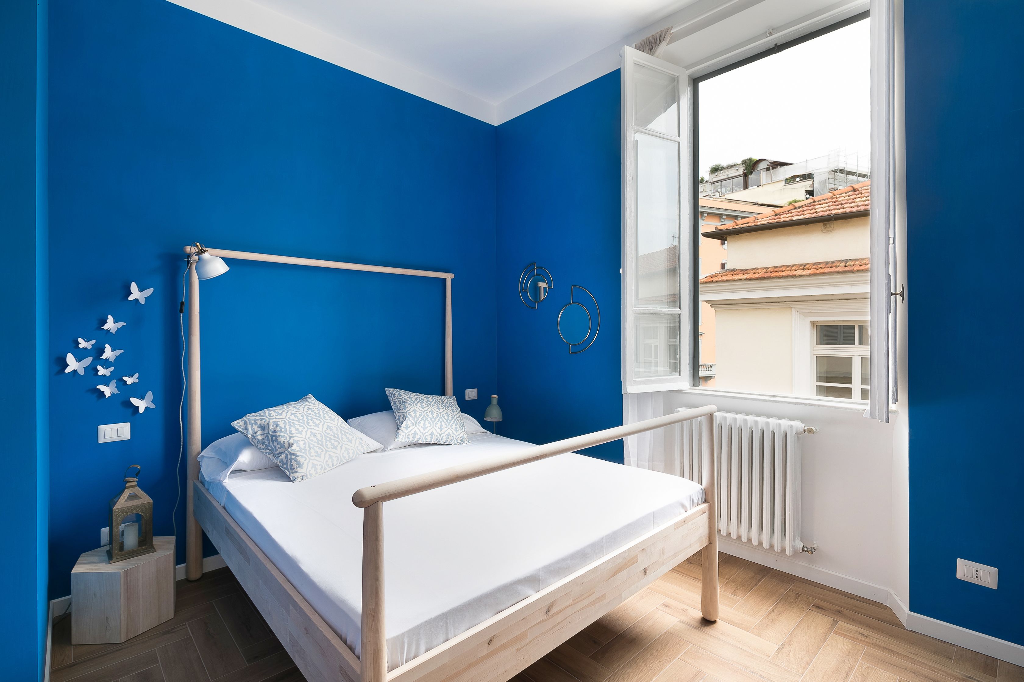 Un dormitorio con cama, ventana, radiador y vistas a edificios.