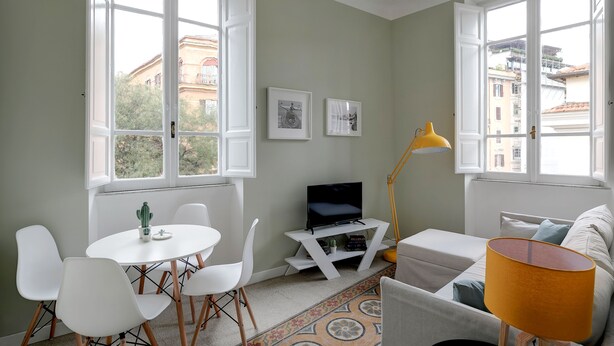 Una sala de estar moderna con un sofá blanco, una mesita redonda con sillas, un televisor sobre un mueble blanco y una lámpara de pie amarilla.