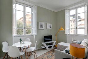 A modern living room with a white sofa, a small round table with chairs, a TV on a white stand, and a yellow floor lamp. - Vatican Sweet Suite (Rome)