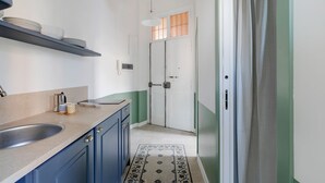 Cocina estrecha con un lavamanos, armarios y una encimera. Se observa una puerta con ventanal y cortina.