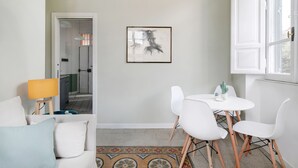 Una sala de estar moderna con un sofá blanco, una mesita con sillas, un cuadro abstracto enmarcado y una ventana con contraventanas blancas.