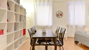 Una habitación con una mesa de comedor, sillas, una estantería, una cama y una ventana con cortinas.