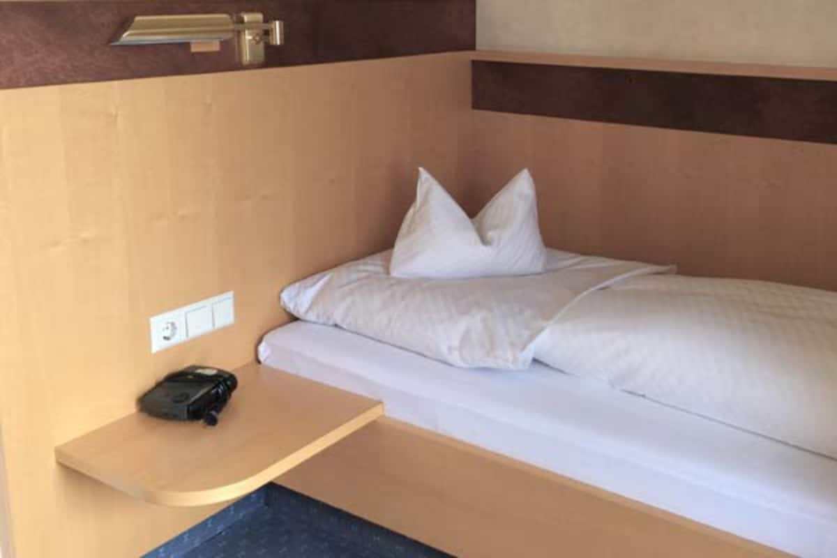 A single bed with white bedding and pillows, a bedside table with a black object, and a wooden headboard.