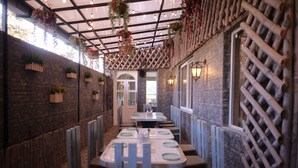 A restaurant with a rustic brick wall, hanging plants, and a wooden lattice on one side. Tables are set with white plates and dark chairs. - Raja ka bagh - A Boutique hotel (Nurpur)