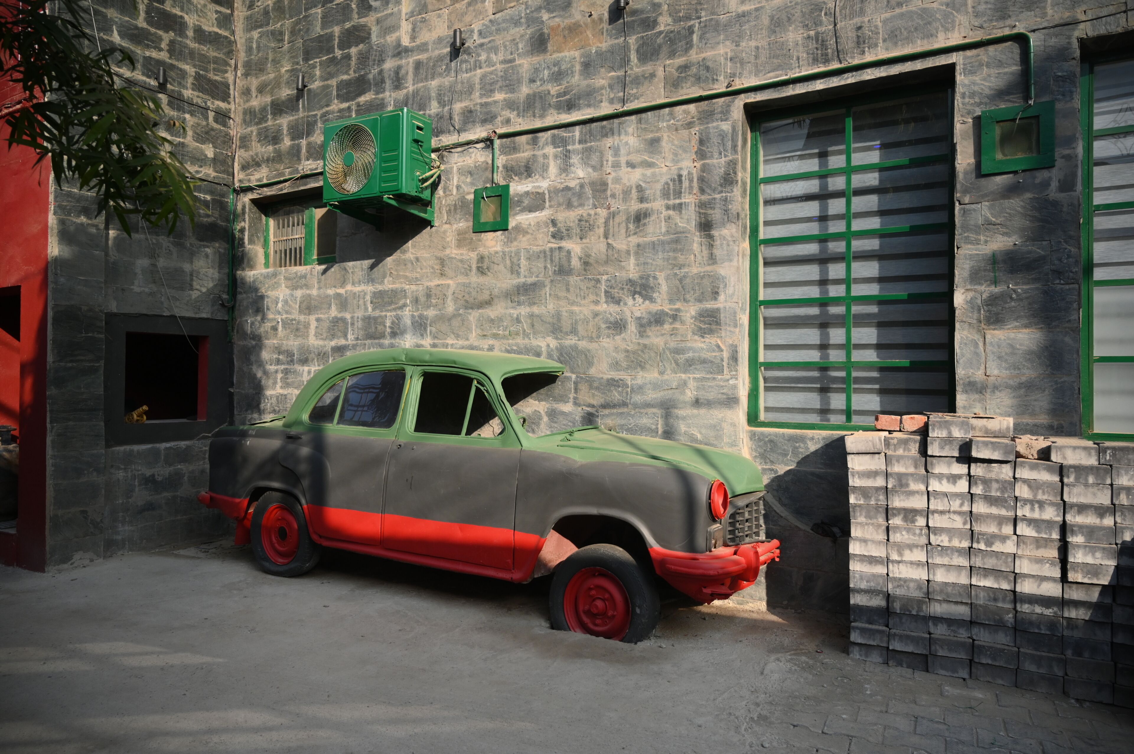 A vintage car parked in front of a stone building with green shutters and a stack of bricks.