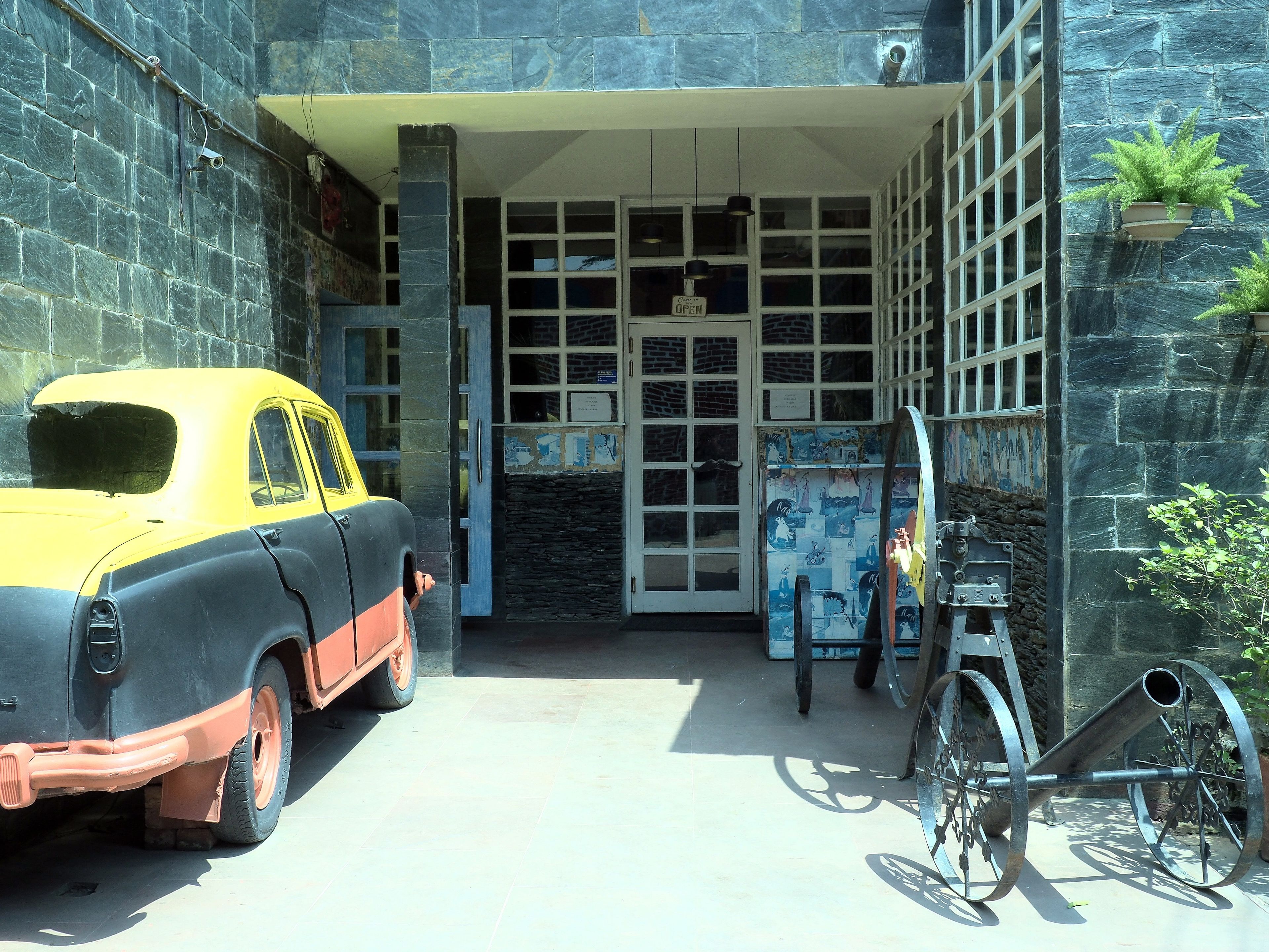 Ein Oldtimer parkt vor einem Gebäude mit einer steinernen Fassade, neben einem Fahrrad, und ein Schild zeigt, dass die Einrichtung geöffnet ist.