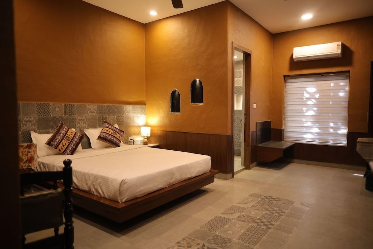 Habitación de hotel con una cama grande, un área de descanso con una silla, una mesita y una ventana con persianas.