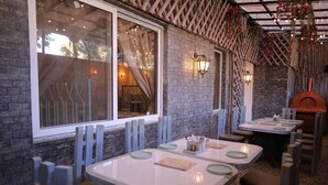 A restaurant with brick walls, large windows, and hanging plants. Tables are set with plates and napkins. - Raja ka bagh - A Boutique hotel (Nurpur)