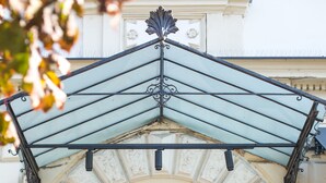 Uma entrada imponente com tapete vermelho, porta de madeira escura e um dossel metálico decorativo.