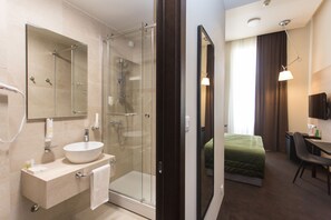 A hotel bathroom with a glass-enclosed shower, a sink with a mirror, and a bed with a green blanket. - Hotel Grand Hedonist (Pancevo)