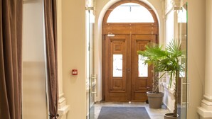 Um saguão de hotel com uma porta dupla de madeira, uma palmeira em vaso e uma porta de vidro à direita.